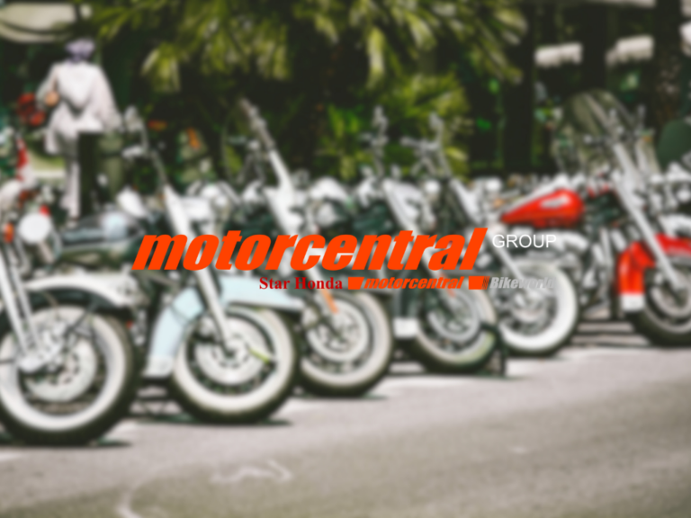 A blurred image of a row of motorcycles parked outdoors. The logo for "motorcentral GROUP" is prominently displayed in bold orange text across the center. Below it, "Star Honda" and "Bike.world" are noted in smaller font, suggesting brand affiliations. The background includes greenery, enhancing the vibrant setting.