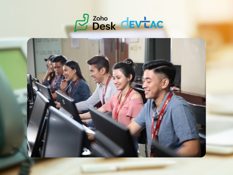 A classroom setting where five students are engaged with computers, smiling and interacting with each other. They are seated in front of desktop monitors, wearing lanyards around their necks. The focus is on a woman in a pink top, who looks happy while speaking to a male colleague next to her. The background features a softly blurred classroom, and the graphic includes the logos for "Zoho Desk" and "DevTac" at the top. The overall atmosphere conveys collaboration and positive engagement in an educational context.