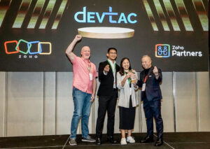 A group photo of four individuals celebrating on stage at the Zoho Inspire Malaysia 2024 event. The backdrop displays the DevTac logo prominently. On the left, a man with short hair in a pink patterned shirt raises his fist in excitement. Next to him, a man in a black suit smiles while holding a small trophy. A woman in a gray blazer stands next to him, proudly holding a trophy. On the right, another man in a black jacket gives a thumbs-up. The atmosphere conveys joy and achievement as they are recognized as the top revenue generator.