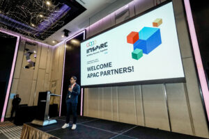 A man in a suit giving a presentation on a stage in a conference room, with a large screen displaying 'WELCOME APAC PARTNERS!' and colorful geometric shapes