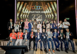A group photo of Zoho partners at the APAC Partner Awards during the Zoho Inspire Malaysia 2024 event. The stage backdrop features the text 'APAC PARTNER AWARDS 2024.' The group consists of diverse individuals, some holding awards, all smiling and posing with thumbs up. They are dressed in professional attire, with a variety of colorful shirts and blazers. The atmosphere is celebratory, highlighting collaboration and achievement among the partners.