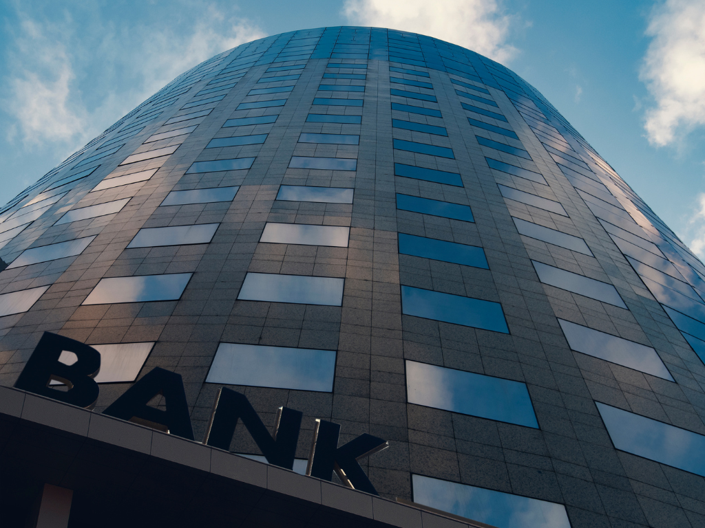 Low-angle view of a tall modern office building with reflective blue-tinted windows. The facade features gray stone panels and a curved, cylindrical shape. The word ‘BANK’ is visible near the entrance at the bottom left.