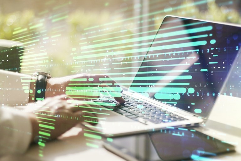 Close-up of hands typing on a laptop with a translucent overlay of digital data streams and neon green/blue lines across the screen, suggesting artificial intelligence or data visualization in a tech workspace.