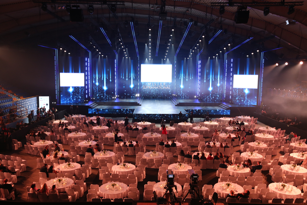 What is Zoho Event Management? A Quick Guide to Organizing Events Event hall with a large stage and bright blue lighting. A grand setup features multiple LED screens and a central illuminated stage, framed by vertical light bars. The audience area is arranged with numerous round tables covered in white cloth and chairs, all facing the stage. There are some people walking and a few cameras or equipment in the foreground, suggesting a formal event or conference.