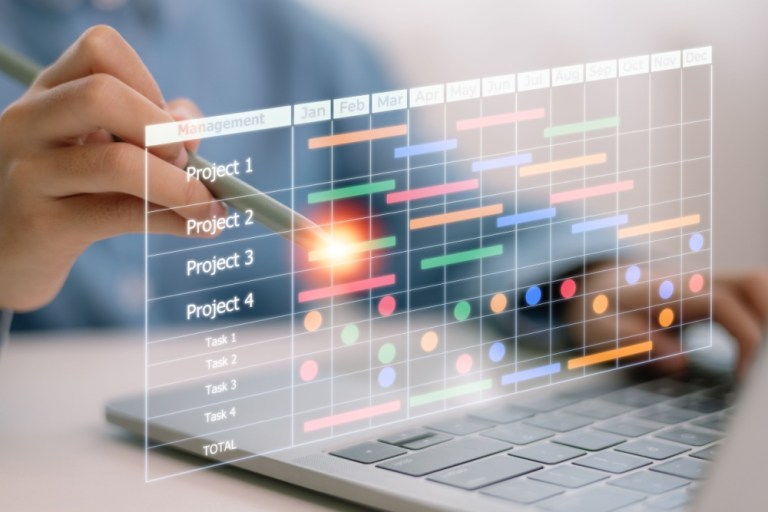 Close-up of a hand drawing or manipulating a translucent holographic project timeline over a laptop. The holographic display shows a grid calendar with rows labeled 'Project 1' through 'Project 4' and 'Task 1' to 'Task 4', color-coded bars and dots representing tasks and milestones across months. The overall scene suggests project planning or scheduling visualization.